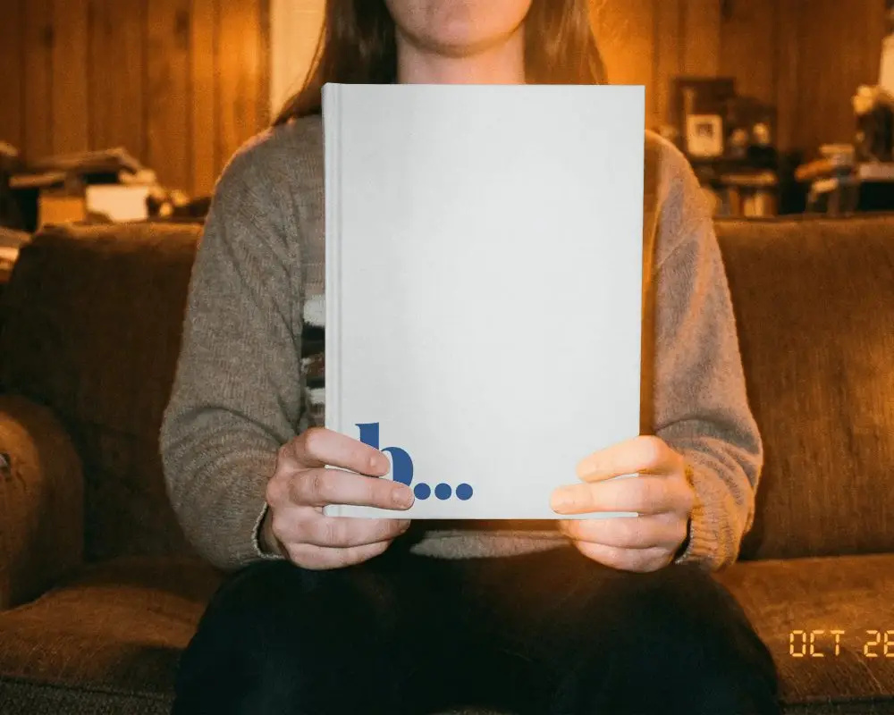 Person Holding Blank Hardcover Book Mockup In Living Room Setting For Custom Designs
