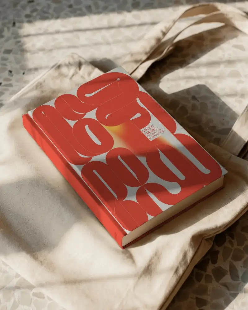 Hardcover Book Mockup Placed On Cream Canvas Tote Bag With Sunlight And Textured Floor Background