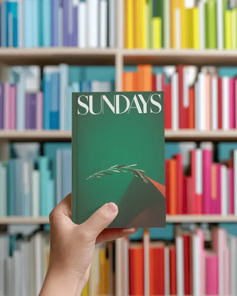 Hand Holding Blank Hardcover Book Mockup Against Colorful Library Bookshelf Background