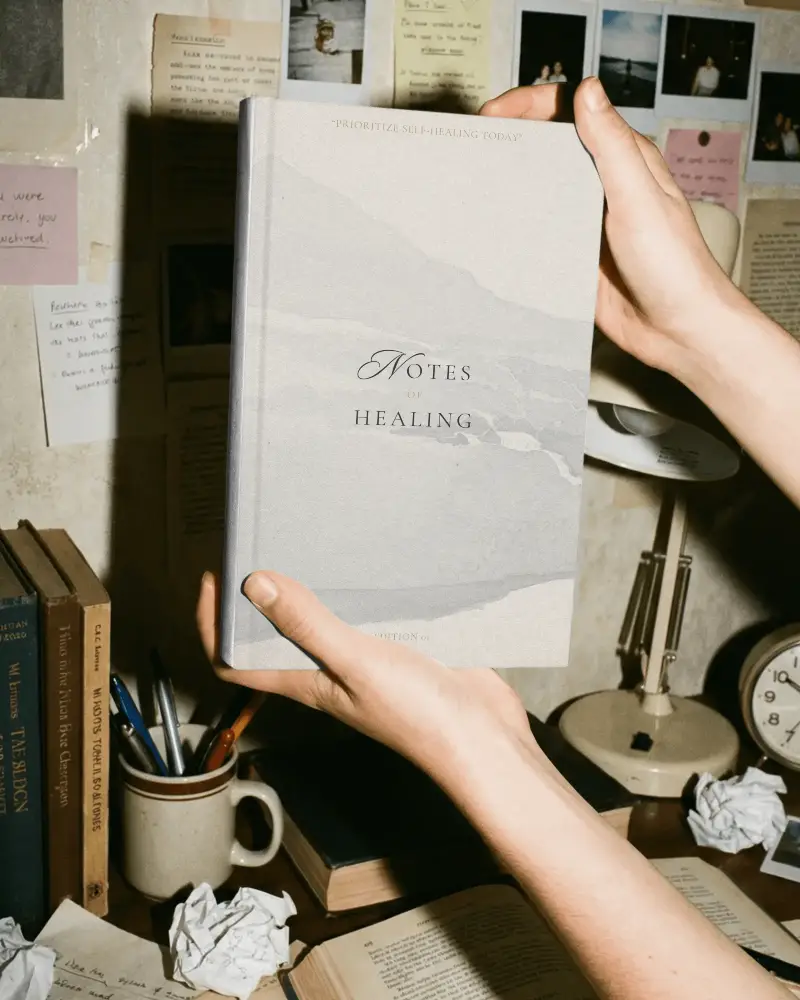 Person Holding Blank Book Mockup On Vintage Styled Desk With Notes And Books