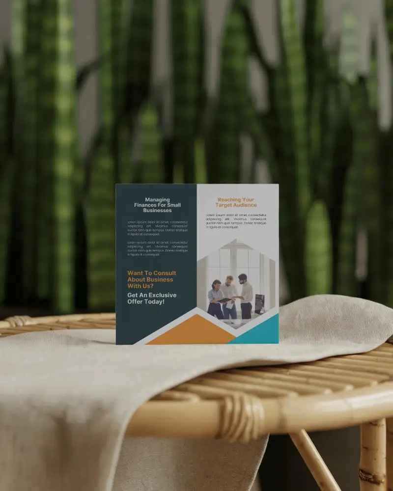 Square Brochure Mockup Standing On Rattan Table With Cloth Cover Against Blurred Background Of Vertical Green Plants