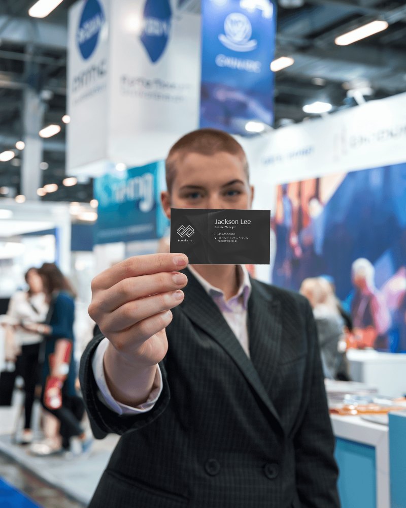 Trade Show Business Card Mockup Focused Hand Holding Card In Lively Exhibition Booth Background