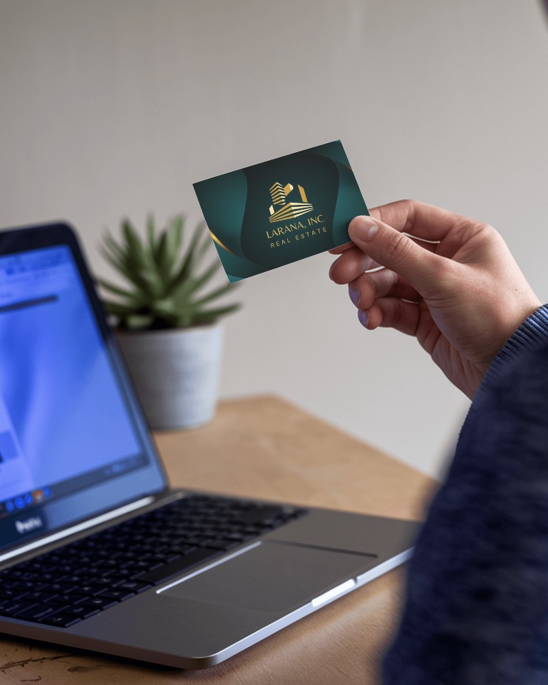 Hand Presenting Business Card Over Laptop On Wooden Table With Plant For Relaxed Work At Home Feel