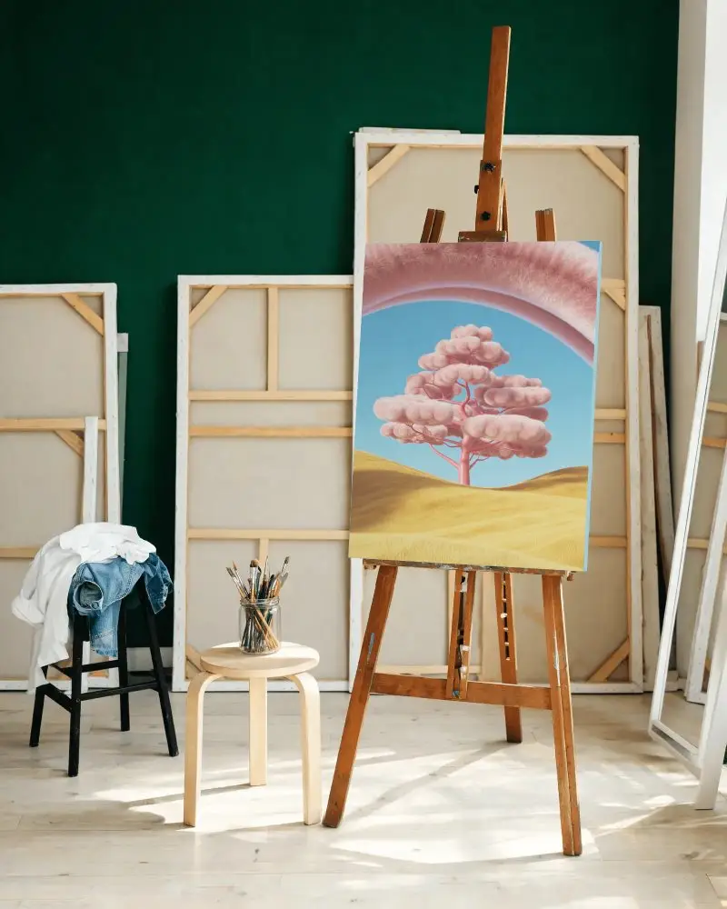 Blank Canvas On Wooden Easel In Artist Studio With Paintbrushes And Frame Backdrop