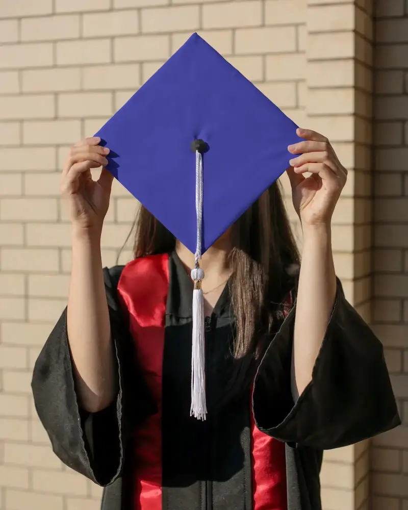 Sunlit Outdoor Graduation Cap Mockup Female Holding Cap Upright In Front Of Face Near Brick Wall