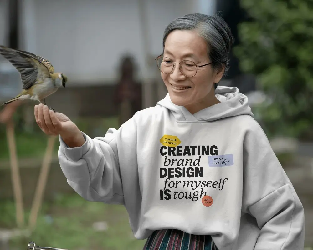 Peaceful Outdoor Hoodie Mockup Elderly Woman Smiling While Feeding Bird In Garden Setting