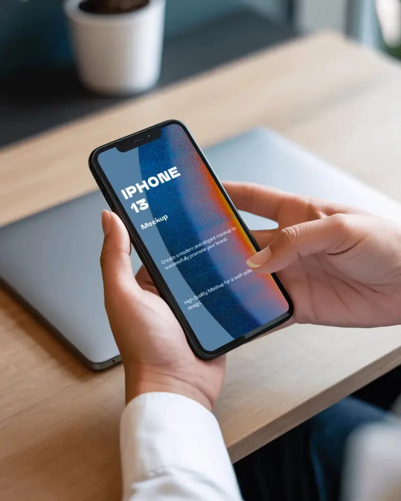 Iphone 13 Mockup In Hands Over Desk With Closed Laptop And Modern Office Background