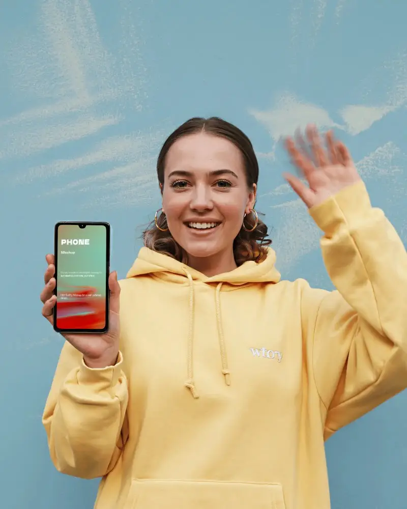 Cheerful Oneplus Smartphone Mockup Smiling Model Holding Phone In Yellow Hoodie Against Blue