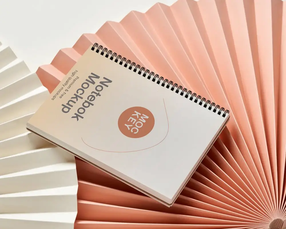 Landscape Spiral Notebook Mockup Resting Diagonally On Layered Accordion Paper Fans