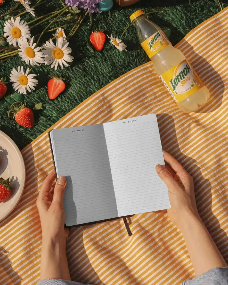 Open Notebook Mockup In Hands On Sunny Picnic Blanket With Flowers And Fresh Summer Vibes