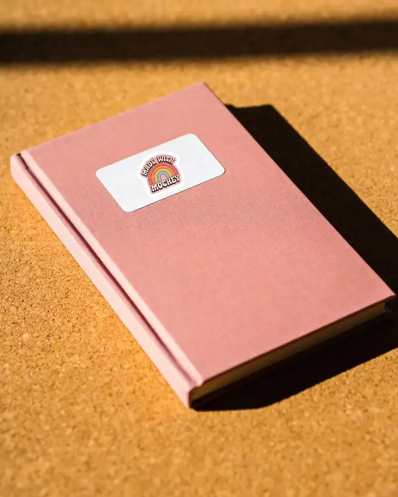 Rectangular Sticker Mockup On Textured Cover Book Placed On Wooden Cork Surface Background