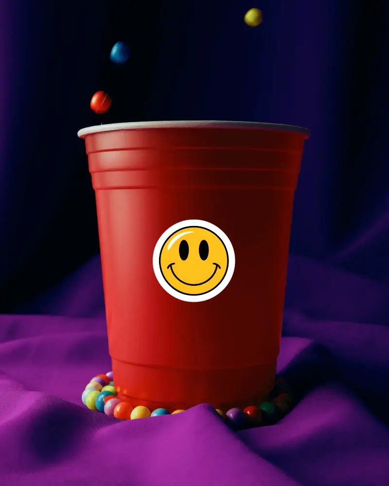 Colorful Candy Beads Surrounding Large Plastic Party Cup Sticker Mockup On Purple Satin Fabric