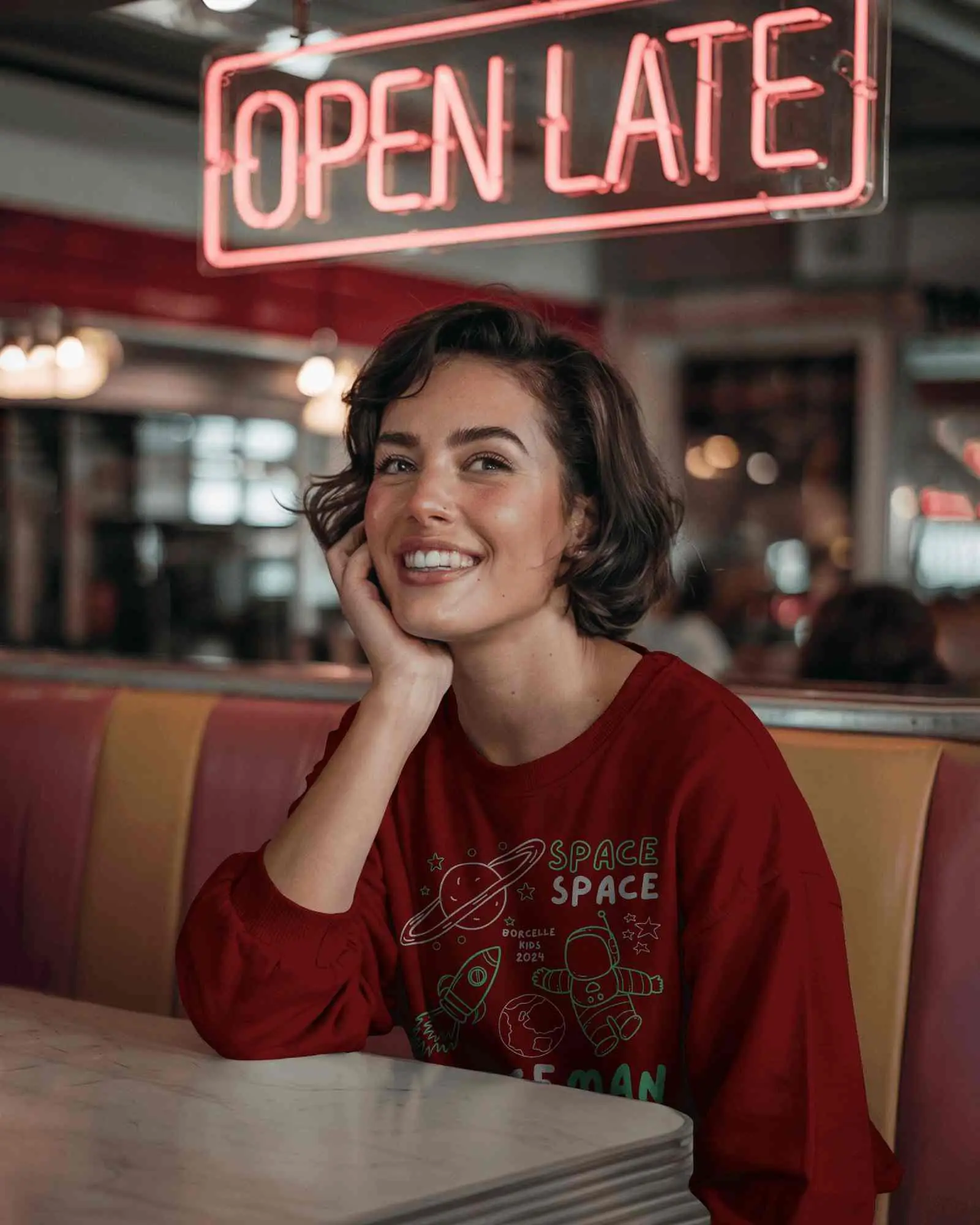 Retro Diner Sweatshirt Mockup Smiling Female Model Sitting In Booth Under Open Late Neon Sign Vibrant And Nostalgic Vibes