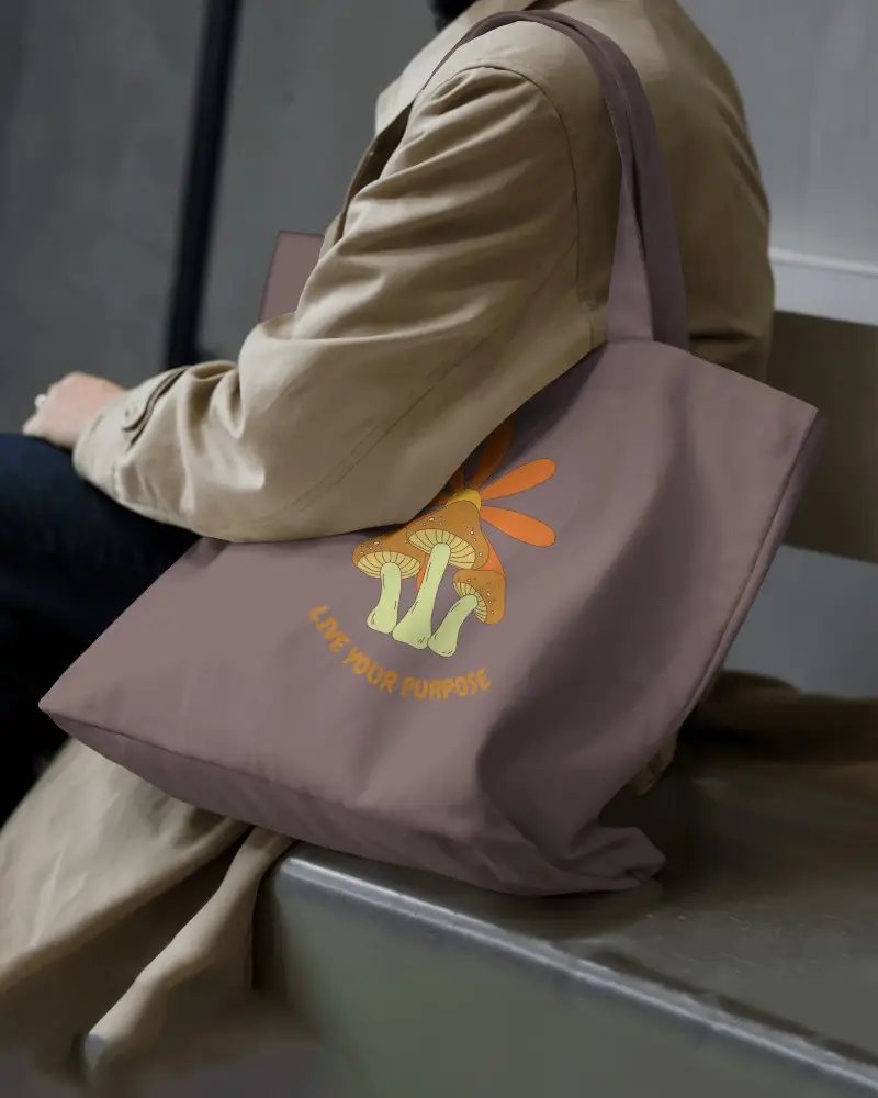 Earthy Toned Totebag Mockup Closeup Side View Seated Male Model In Trench Coat Casual Urban Setting Illustrated Mushroom Design Live Your Purpose Text Retro Style Inspiration