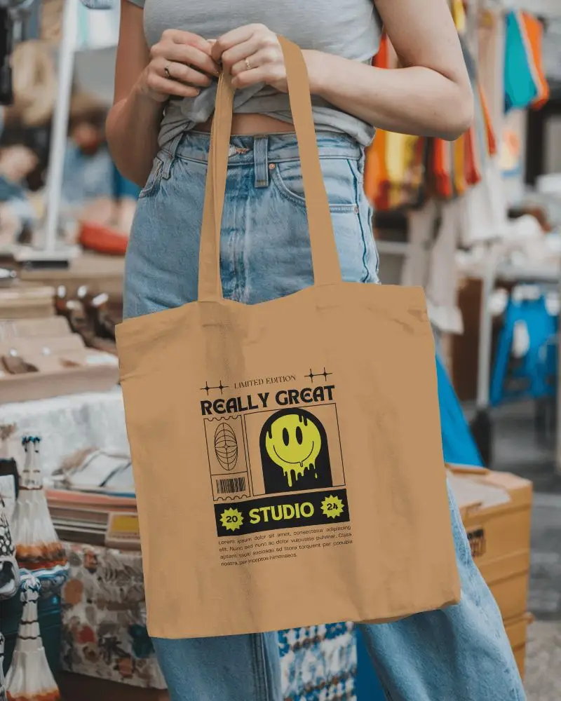 Street Style Totebag Mockup Basic Tote Highlighting Design Space Carried By A Young Woman Dressed In Jeans And A Crop Top In Front Of A Colorful Market Backdrop Fashionable And Authentic Atmosphere