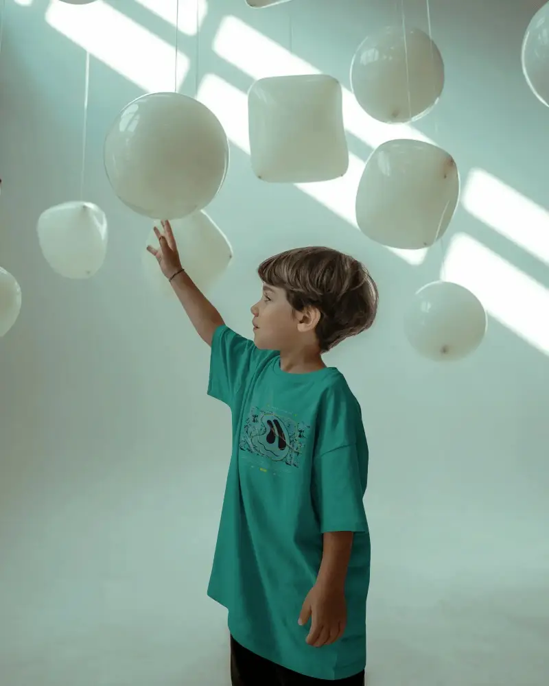 Kids Relaxed Fit Tshirt Mockup Standing Indoor With Floating Balloons For Design Presentation
