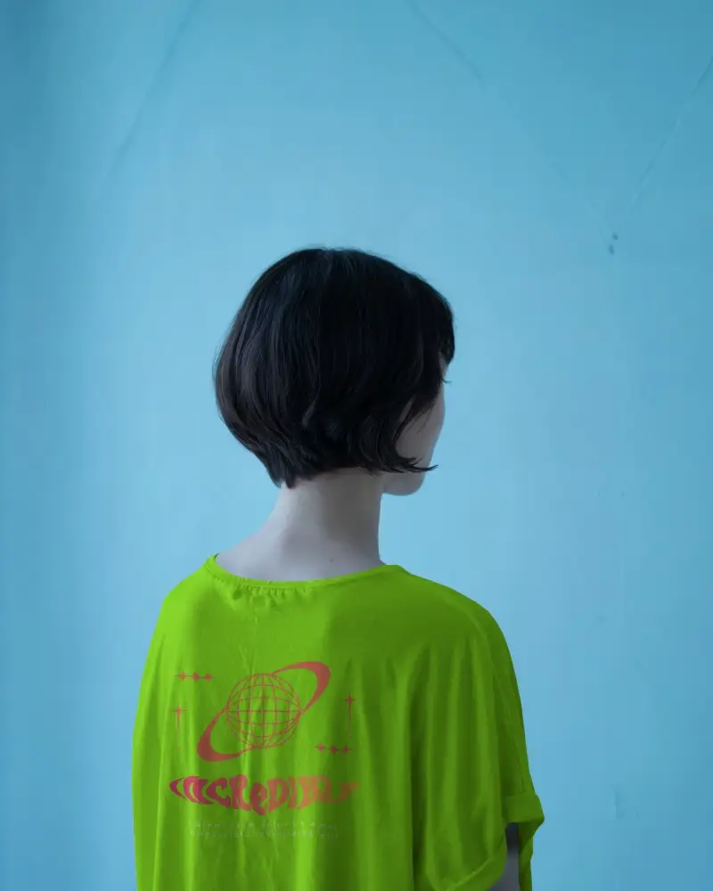 Back View Tshirt Mockup Female Model Dark Bob Cut Light Beige Shirt Against Blue Wall
