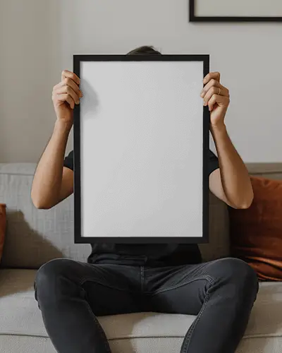 Blank Poster Mockup Held By Seated Person On Couch For Artwork Presentation