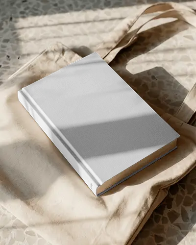 Hardcover Book Mockup Placed On Cream Canvas Tote Bag With Sunlight And Textured Floor Background