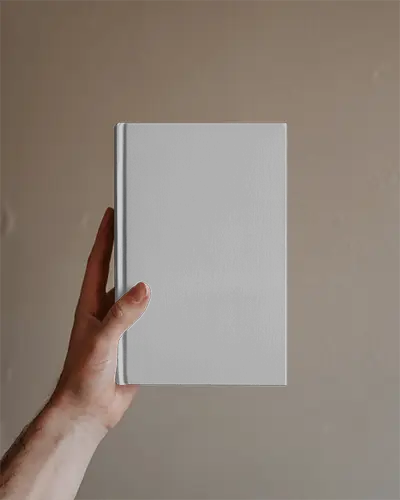 Blank Hardcover Book Mockup Held Upright Against Muted Brown Wall Closeup Of Hand And Simple Setting
