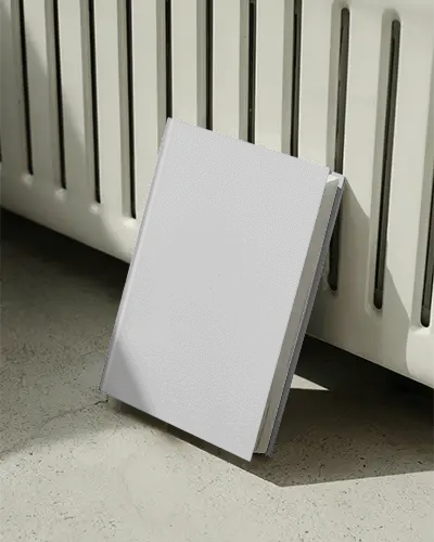 Hardcover Book Mockup Standing Against Radiator In Natural Light On Textured Floor