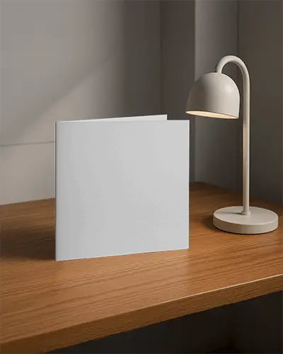Square Folded Brochure Mockup Standing On Wooden Table Next To Modern Desk Lamp Soft Interior Lighting Cozy Minimal Scene Clean Promotional Display Setup