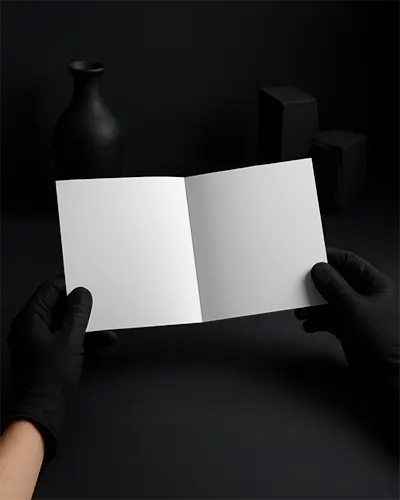 Square Brochure Mockup Gloved Hands Holding Open Bifold On Dark Minimalist Surface High Contrast Shadow Presentation