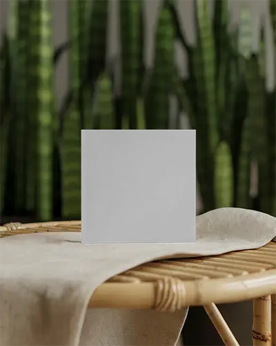 Square Brochure Mockup Standing On Rattan Table With Cloth Cover Against Blurred Background Of Vertical Green Plants