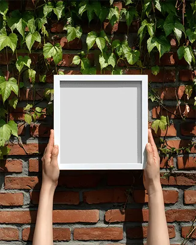 Hands Holding Square Photo Frame Mockup Against Brick Wall With Green Trailing Vines Background