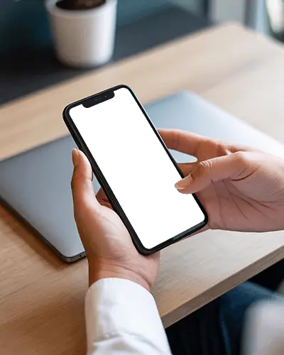 Iphone 13 Mockup In Hands Over Desk With Closed Laptop And Modern Office Background