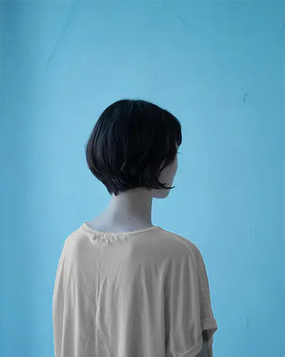 Back View Tshirt Mockup Female Model Dark Bob Cut Light Beige Shirt Against Blue Wall