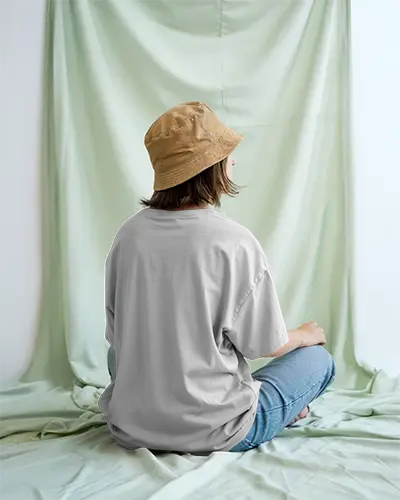 Relaxed Backdrop T Shirt Mockup Female Model Sitting Cross Legged In Light Green Drape Studio Wearing Bucket Hat Natural Lighting Boho Vibe