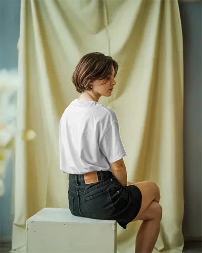 Tshirt Mockup Soft Curtain Background Female Model Seated On Box Looking Away Showing Back Design Subtle Lighting Studio Style