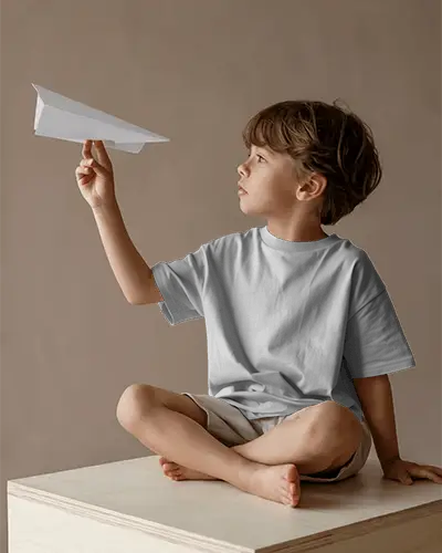 Kid Playing Paper Plane T Shirt Mockup Child Sitting On Wooden Box In Natural Light Setting