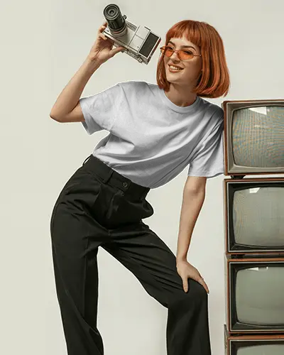 Retro Tshirt Mockup Female Model With Orange Glasses Holding Camera Beside Vintage Tvs