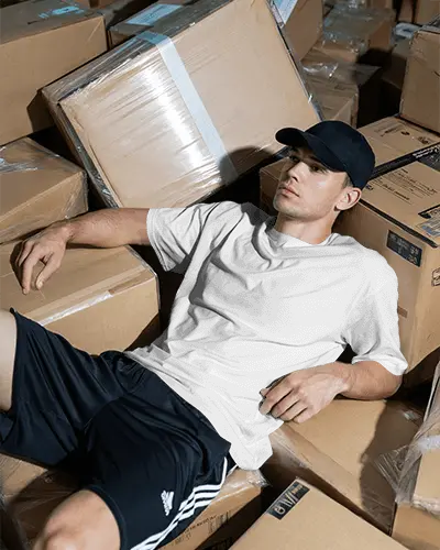 Warehouse Style T Shirt Mockup Male Model Relaxing Among Cardboard Boxes Casual Look