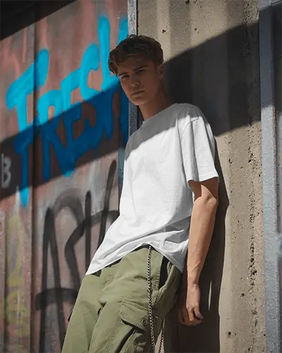 Urban Street Style T Shirt Mockup Worn By Young Man Leaning Against Rough Wall Background