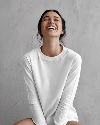 Joyful Studio Blank Round Neck Long Sleeve T Shirt Mockup Woman In White Tee Laughing Against Textured Gray Background 0831