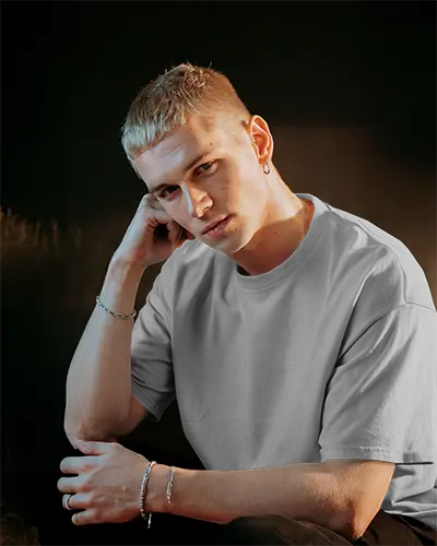 Moody Indoor T Shirt Mockup Blonde Male Model Sitting In Soft Shadowed Lighting Relaxed And Contemporary Vibe