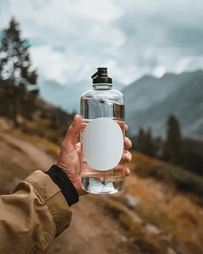 Personalized Sticker Mockup On Clear Water Bottle Held In Hand Mountain Trail Setting