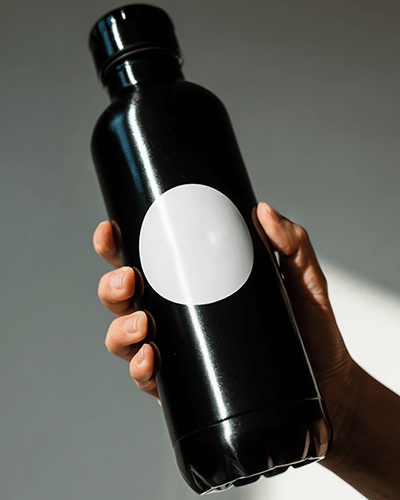 Hand Holding Black Bottle Sticker Mockup With Glossy Surface And Round Label Under Directional Light 0273