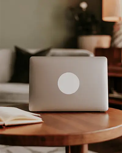 Sleek Laptop With Round Sticker Mockup On Backside Placed On Wooden Table In Cozy Room Setting
