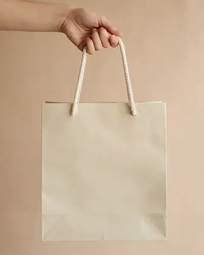 Neutral Paper Bag Sticker Mockup With Hand Holding Rope Handles On Minimal Beige Background