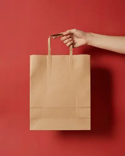 Minimal Kraft Paper Bag Sticker Mockup With Hand Holding On Bold Red Background For Brand Showcase