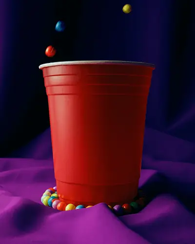 Colorful Candy Beads Surrounding Large Plastic Party Cup Sticker Mockup On Purple Satin Fabric