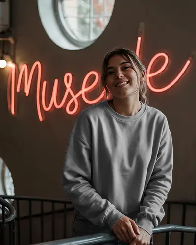 Neon Lit Indoor Sweatshirt Mockup Female Model Smiling Wearing Grey Crewneck By Railings Modern Artsy Vibe With Muse Sign
