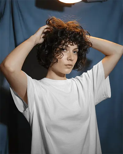 Moody Studio T Shirt Mockup Curly Haired Female Model In Overhead Lighting Against Blue Backdrop