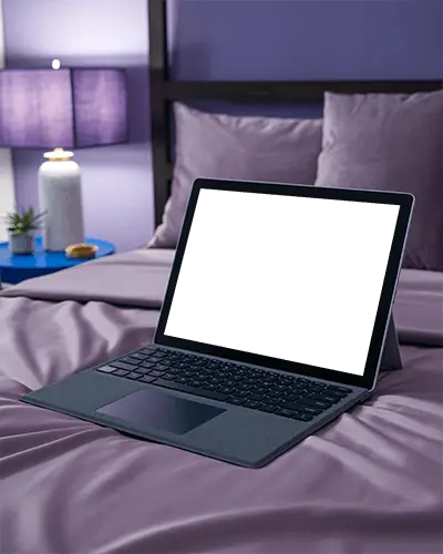 Microsoft Surface Laptop Mockup On Bed In Cozy Bedroom With Purple Sheets And Warm Night Lamp Light
